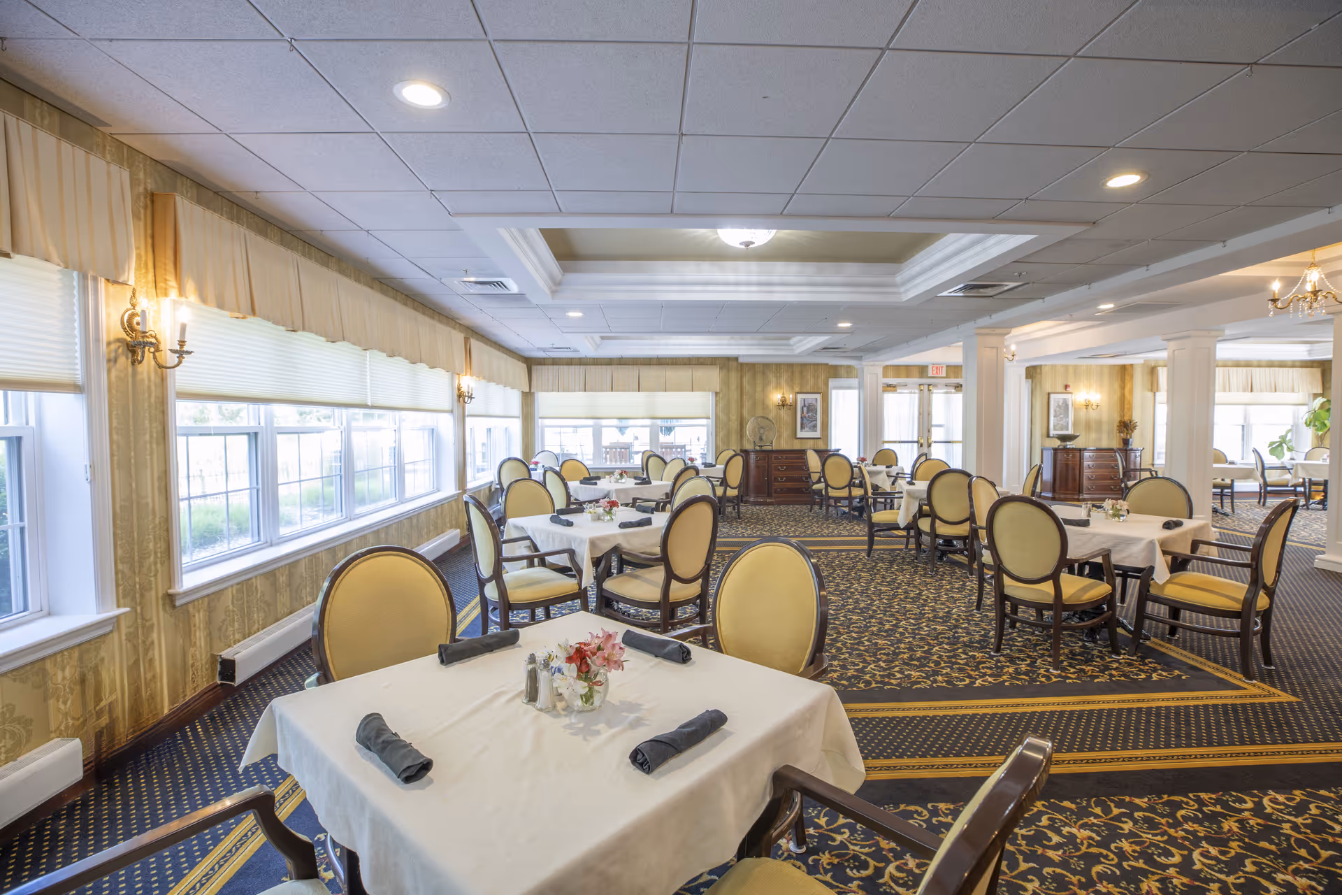 A spacious dining room with multiple tables covered in white tablecloths, each set with rolled black napkins and small floral centerpieces. The room features large windows with beige valances, patterned carpet, and elegant wall sconces providing warm lighting. The ceiling has recessed lighting and a decorative tray ceiling in the center.