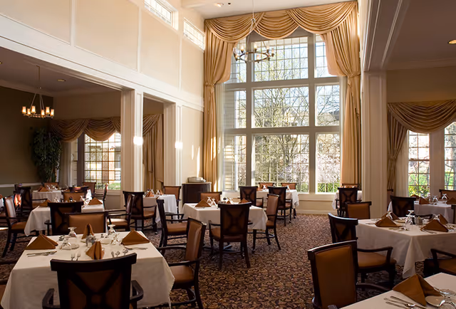 A spacious dining room with multiple tables covered in white tablecloths and set with brown napkins, glasses, and silverware. The room features large windows with elegant drapes, allowing natural light to fill the space. There are several wooden chairs around each table, and a chandelier hangs from the ceiling.