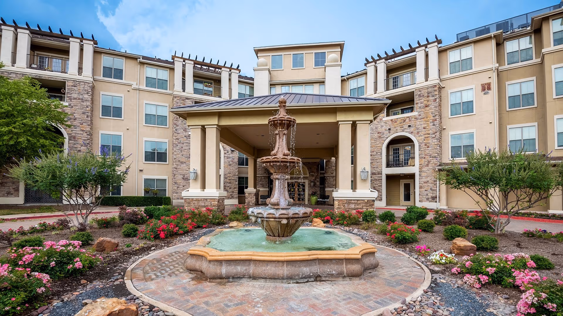 Front exterior view of a senior living facility named Atria Canyon Creek featuring a large covered entrance with a tiered water fountain in the center, surrounded by landscaped flower beds and shrubs.