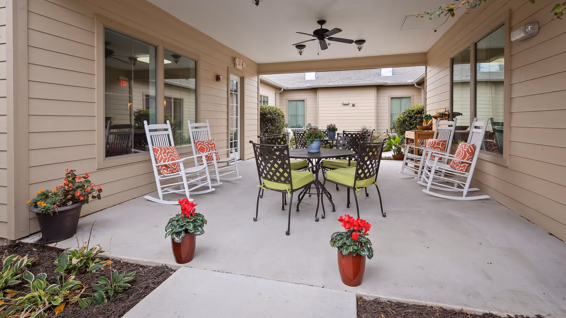 Covered outdoor patio area with beige siding walls, featuring four white rocking chairs with orange patterned cushions, a round black metal table with four green cushioned chairs, and several potted plants including red flowers in the foreground.