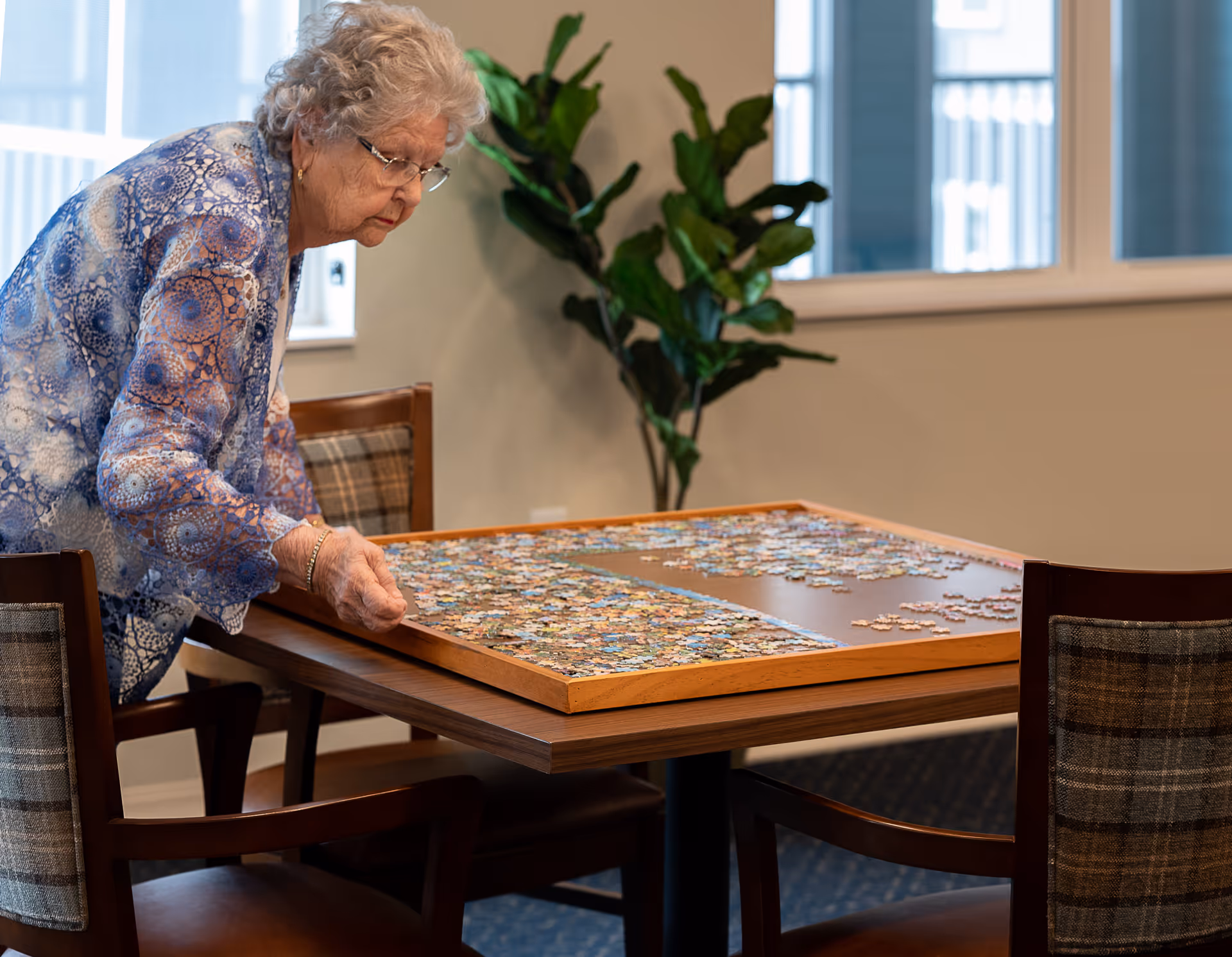 An elderly woman with gray hair and glasses is standing at a wooden table working on a large jigsaw puzzle. The table is surrounded by wooden chairs with plaid cushions. In the background, there is a green leafy plant and large windows letting in natural light.