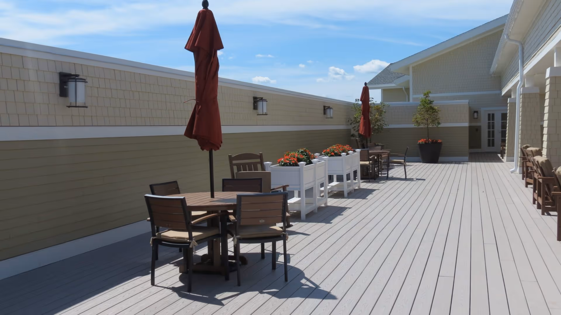 Outdoor patio area with wooden tables and chairs, closed red umbrellas, white planter boxes with red flowers, and potted trees along a beige and light green building wall under a blue sky with some clouds.