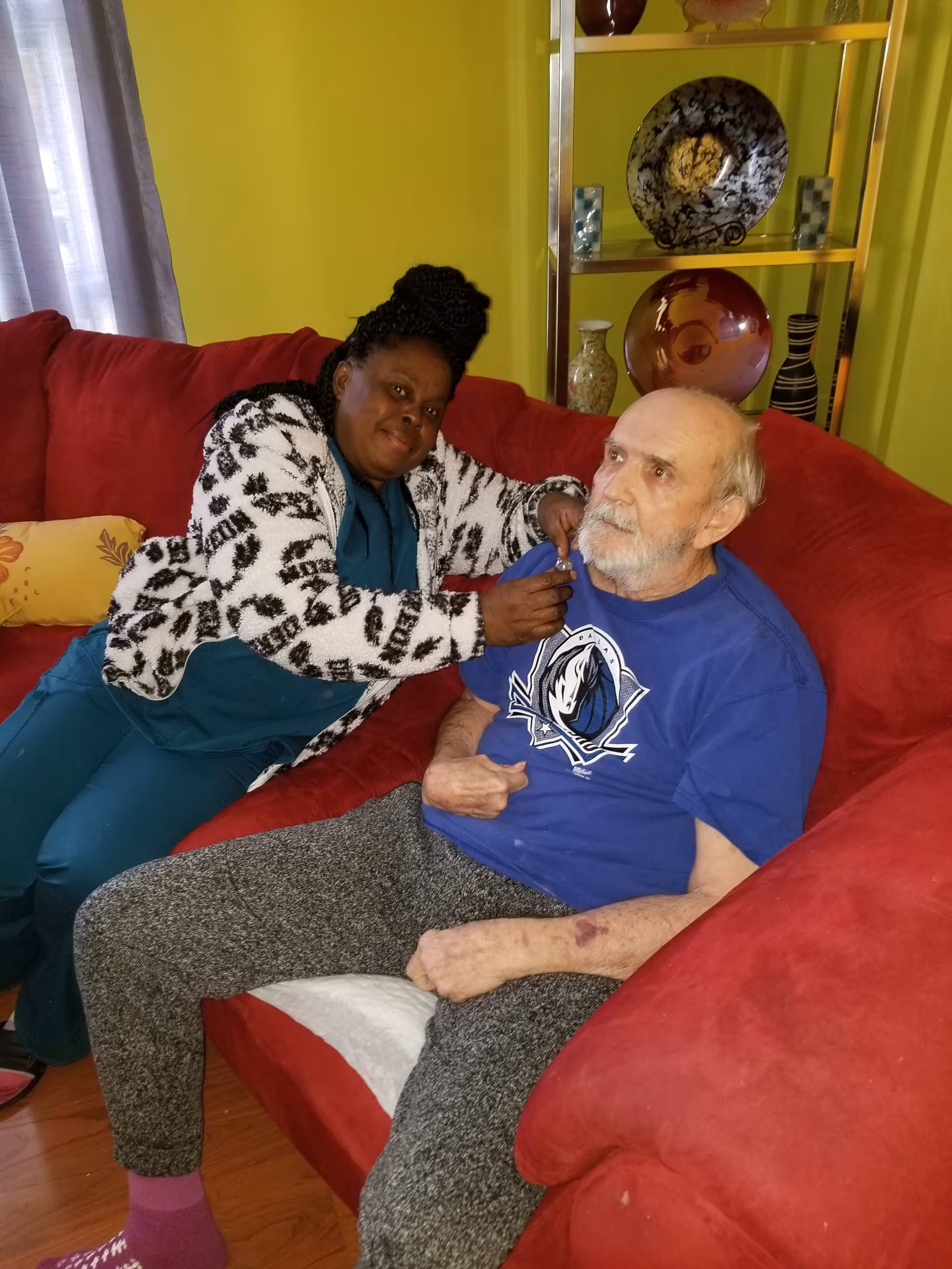 A woman with braided hair wearing a patterned jacket and teal scrubs is sitting on a red couch next to an elderly man with a beard wearing a blue t-shirt and gray sweatpants. The woman is gently holding the man's chin. Behind them is a yellow-green wall with a metal shelf displaying decorative plates and vases.