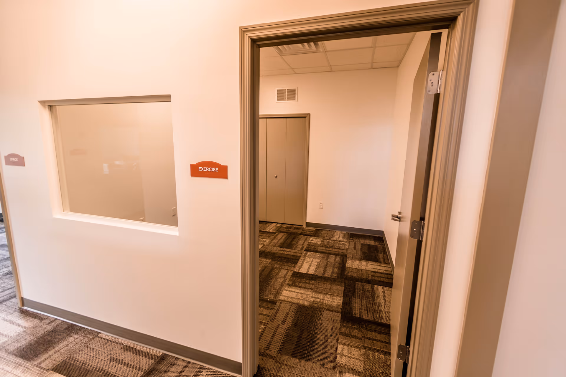 Interior hallway of a senior living facility showing an open door leading to a room with patterned carpet. On the wall outside the door, there is a red sign labeled 'EXERCISE' and a window looking into another room labeled 'OFFICE'.