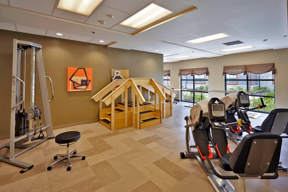 Well-lit fitness room with cardio machines, a cable weight station, a wooden therapy staircase, and large windows.
