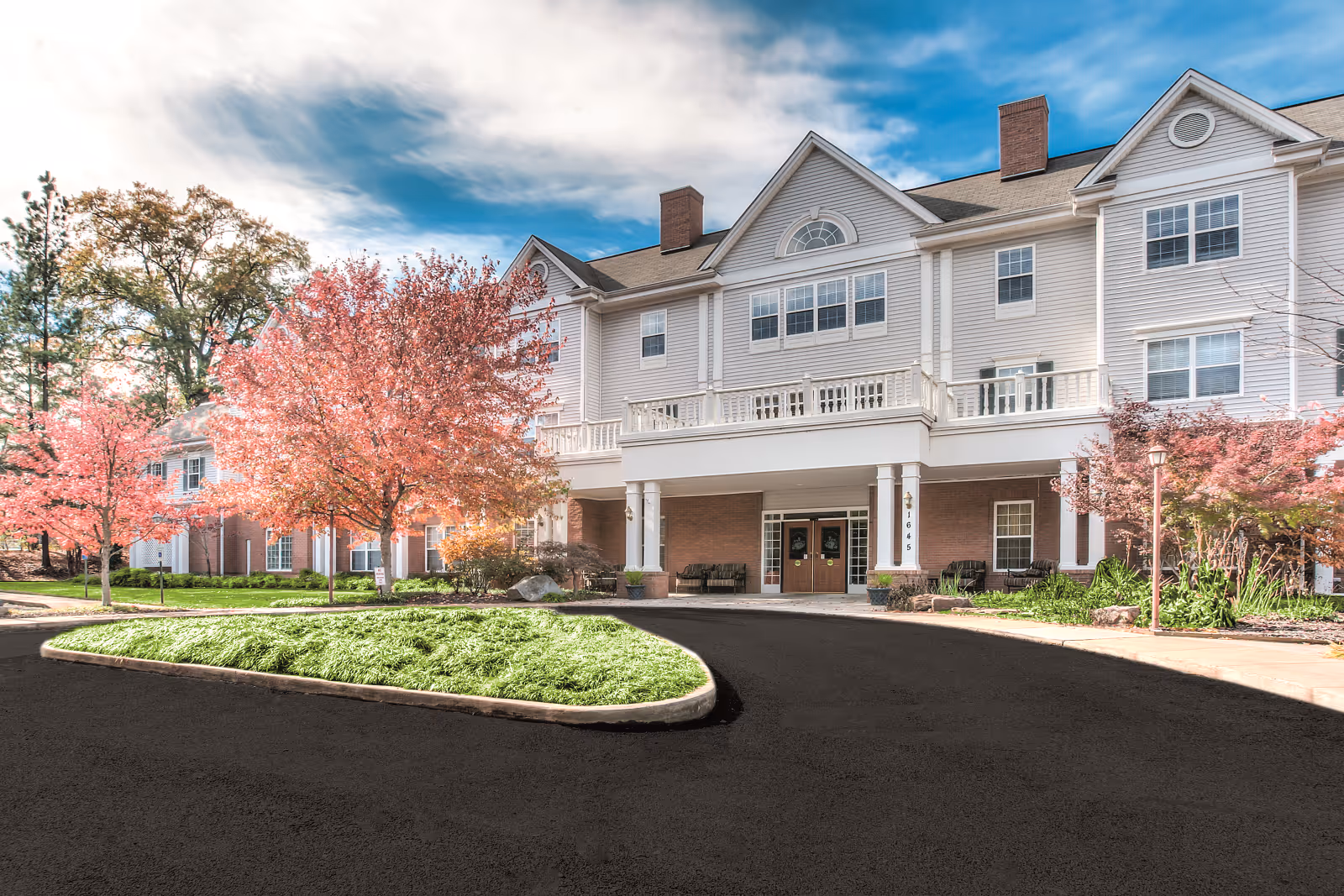 Exterior view of Avaline of River Oaks, a multi-story senior living facility with a curved driveway, landscaped garden beds, trees, and a covered entrance with double doors and seating areas.