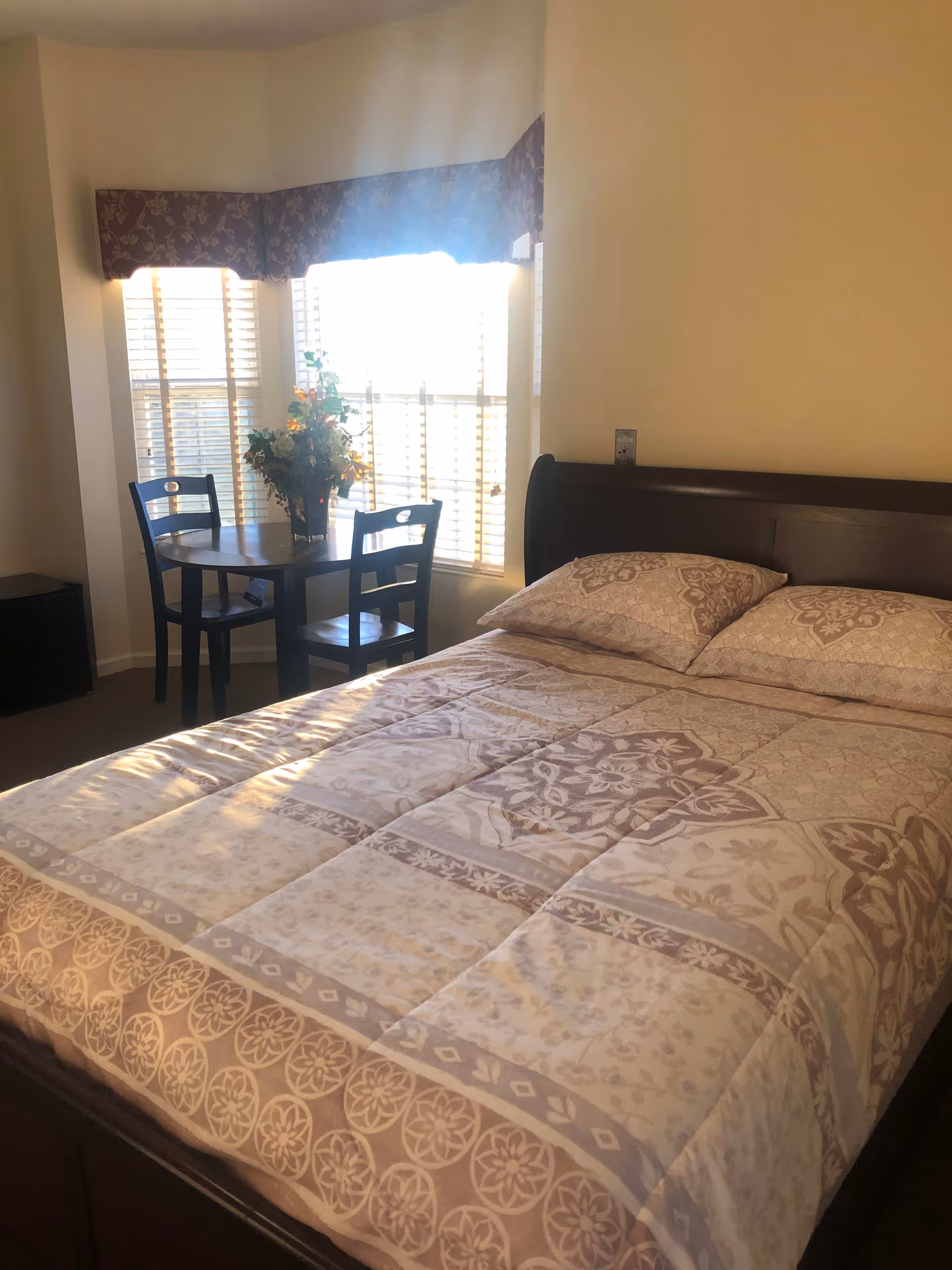 Sunlit bedroom with a made double bed, patterned bedding, and a small round table with two chairs by a window.