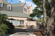 Outdoor patio area at The Courville At Manchester featuring tables with umbrellas, surrounded by trees and landscaping, adjacent to a beige building with green roof shingles and multiple windows.