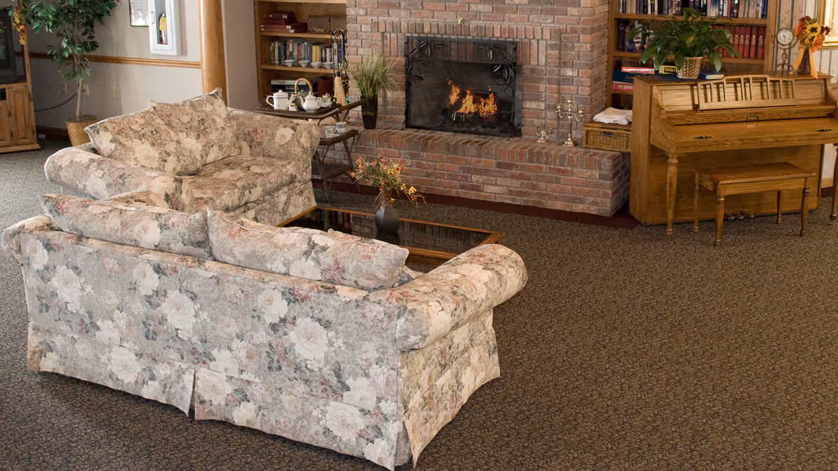 A cozy living room with floral patterned sofas arranged around a glass coffee table, a brick fireplace with a fire burning, wooden bookshelves filled with books, and a wooden piano with a bench. There are plants and decorative items placed around the room.