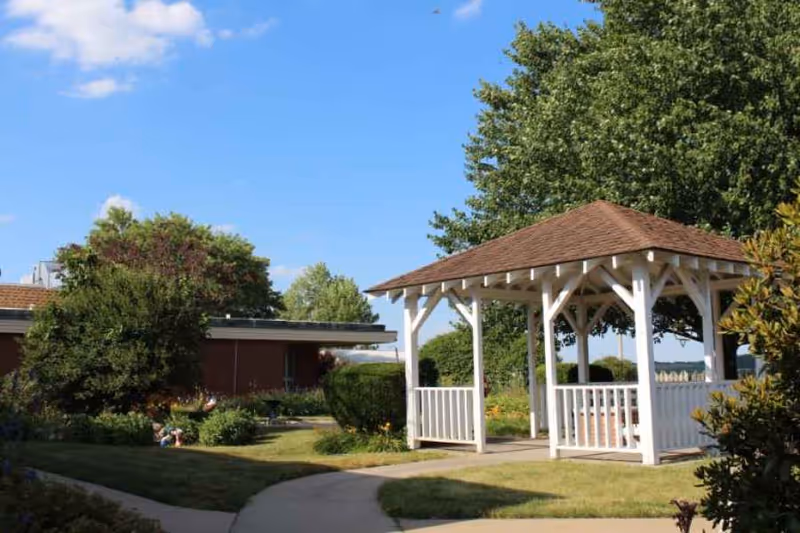 A white wooden gazebo with a brown shingled roof in a garden area with green grass, bushes, and trees under a blue sky with a few clouds. A building is partially visible in the background.
