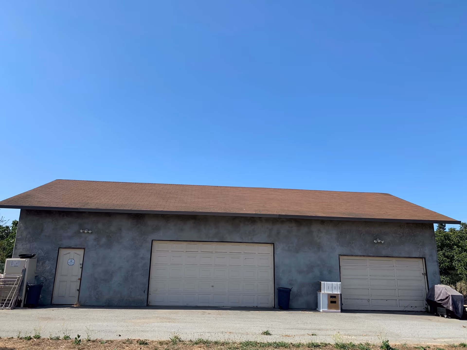 Exterior view of a building with a brown sloped roof and gray walls, featuring two large white garage doors and one smaller white door on the left side. There are some items placed near the doors, including a covered object on the right and a trash bin on the left. The sky is clear and blue.