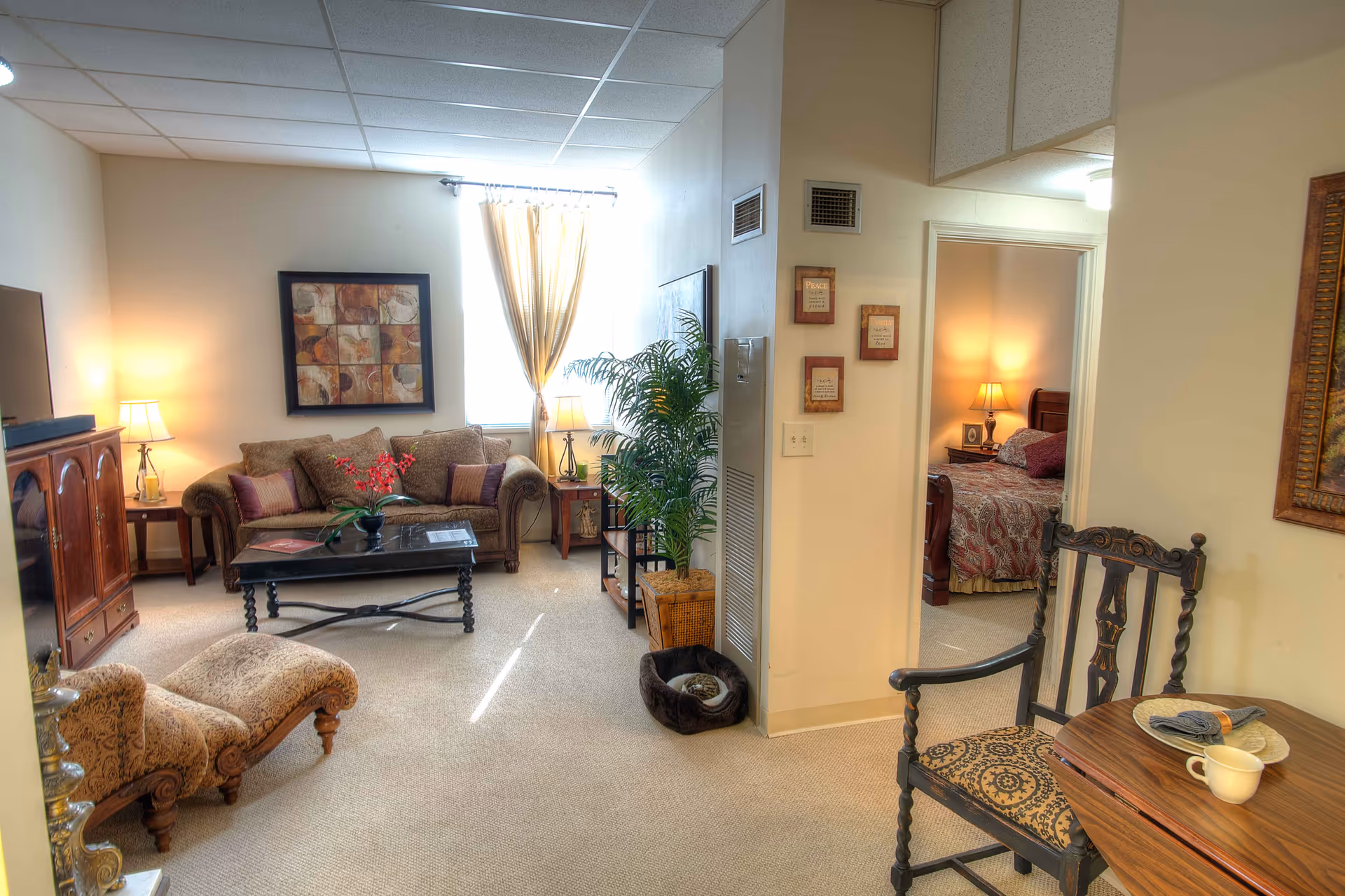 A cozy living room area with a patterned sofa, a coffee table with a flower arrangement, two side tables with lamps, a TV cabinet, and a chaise lounge. There is a window with light-colored curtains letting in natural light. To the right, an open doorway reveals a bedroom with a bed, nightstand, and lamp. A decorative chair and small table with a cup and napkin are in the foreground. The walls have framed artwork and inspirational plaques, and a potted plant is near the doorway.