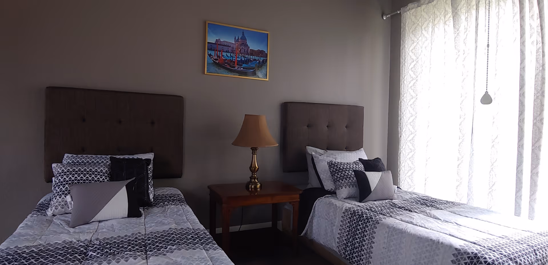 Two twin beds with patterned bedding, a shared nightstand and lamp between them, a framed painting on the wall, and sheer curtains covering a window.