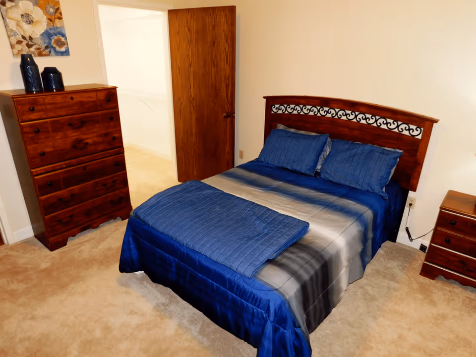 A bedroom with a wooden bed frame featuring decorative metalwork, a bed made with blue and gray bedding, a wooden dresser with two blue vases on top, a wooden nightstand, beige carpet, and an open wooden door leading to another room.