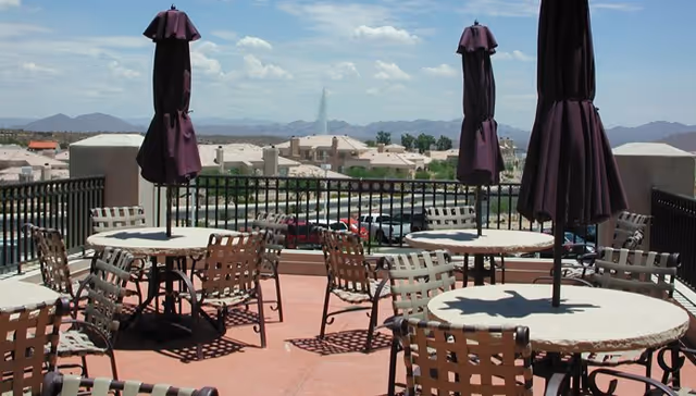 Outdoor patio area with round tables and metal chairs, each table shaded by a closed purple umbrella. In the background, there is a view of rooftops, a water fountain, and distant mountains under a partly cloudy sky.