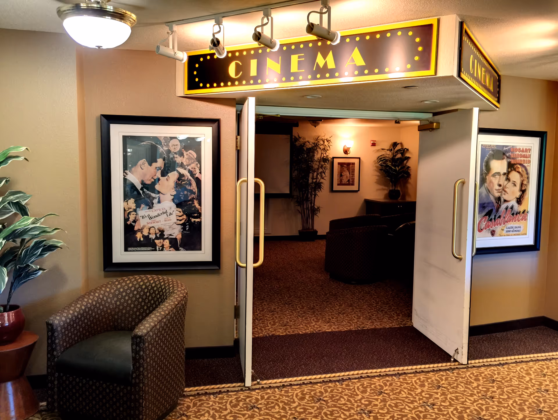 Entrance to a small on-site cinema with an illuminated 'CINEMA' marquee, open double doors, framed movie posters, potted plants and seating.