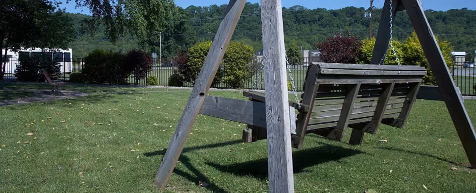 A wooden outdoor swing set with two bench-style swings on a grassy lawn, surrounded by trees and bushes with a fence and hills in the background under a clear sky.