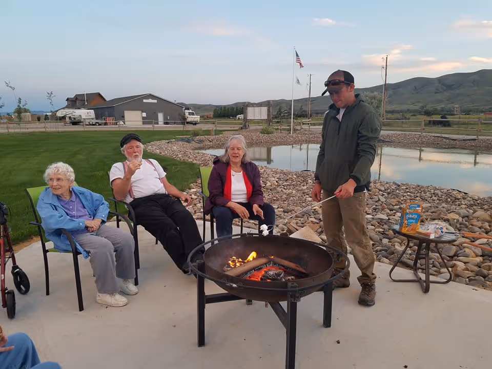 Three elderly people sitting on chairs around a fire pit roasting marshmallows, with a man standing and roasting marshmallows on a stick. They are outdoors near a pond with rocks and grass, with hills and a building in the background under a partly cloudy sky.