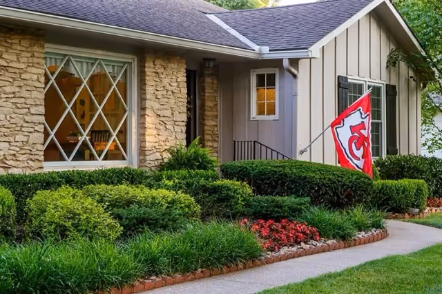 Exterior view of a single-story house with stone and siding walls, a large window with diamond-shaped panes, a Kansas City Chiefs flag mounted near the entrance, and a well-maintained garden with green shrubs and red flowers along a paved walkway.