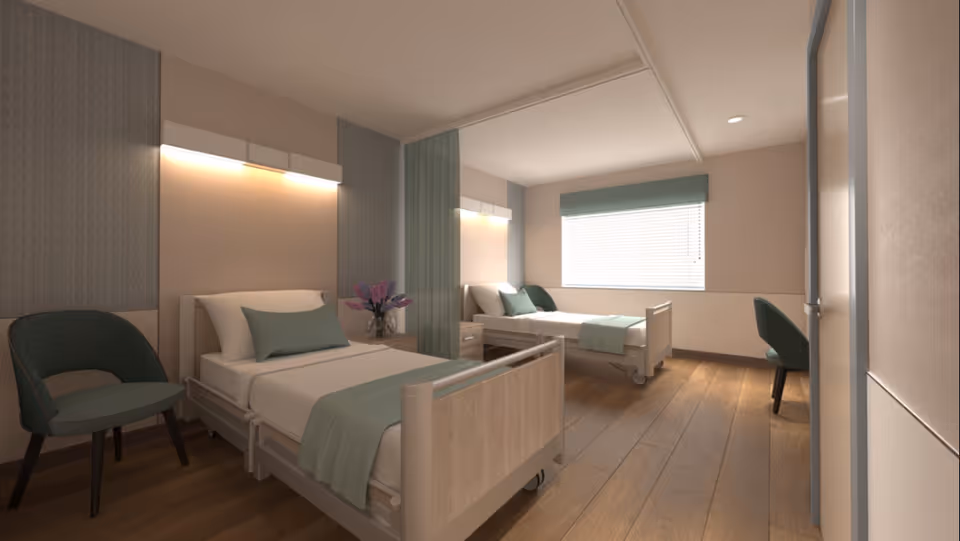 A bright shared patient room with two hospital-style beds, chairs, a nightstand with flowers, and a window with blinds.