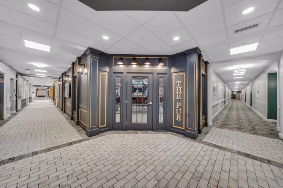 Interior hallway of a senior living facility with a central entrance to a pub labeled 'THE PUB' in gold letters on dark paneling. The hallway has light-colored tiled flooring and white ceilings with recessed lighting, extending to the left and right with doors and wall decorations along the corridors.
