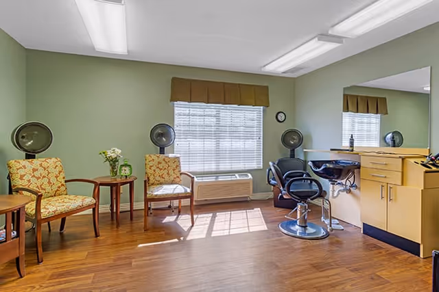 A bright room with wooden flooring featuring two patterned armchairs and a small round table with a vase of flowers near a window with blinds and a brown valance. On the right side, there is a salon styling station with a large mirror, a black salon chair, and hair drying equipment mounted on the wall.
