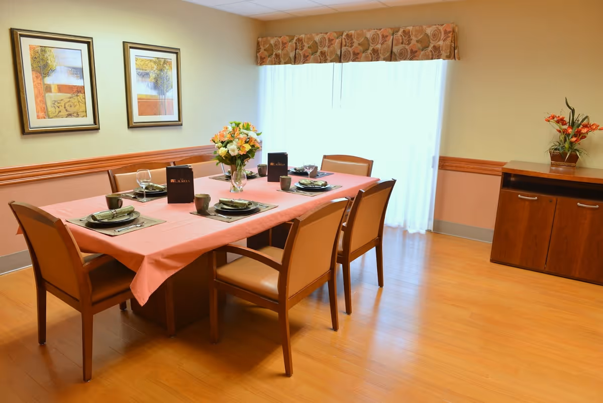 A dining room with a rectangular table covered with a pink tablecloth, set for six people with plates, cups, and glasses. There is a vase with flowers in the center of the table. The room has wooden flooring, two framed paintings on the wall, a window with sheer curtains and a floral valance, and a wooden cabinet with a flower arrangement on top.