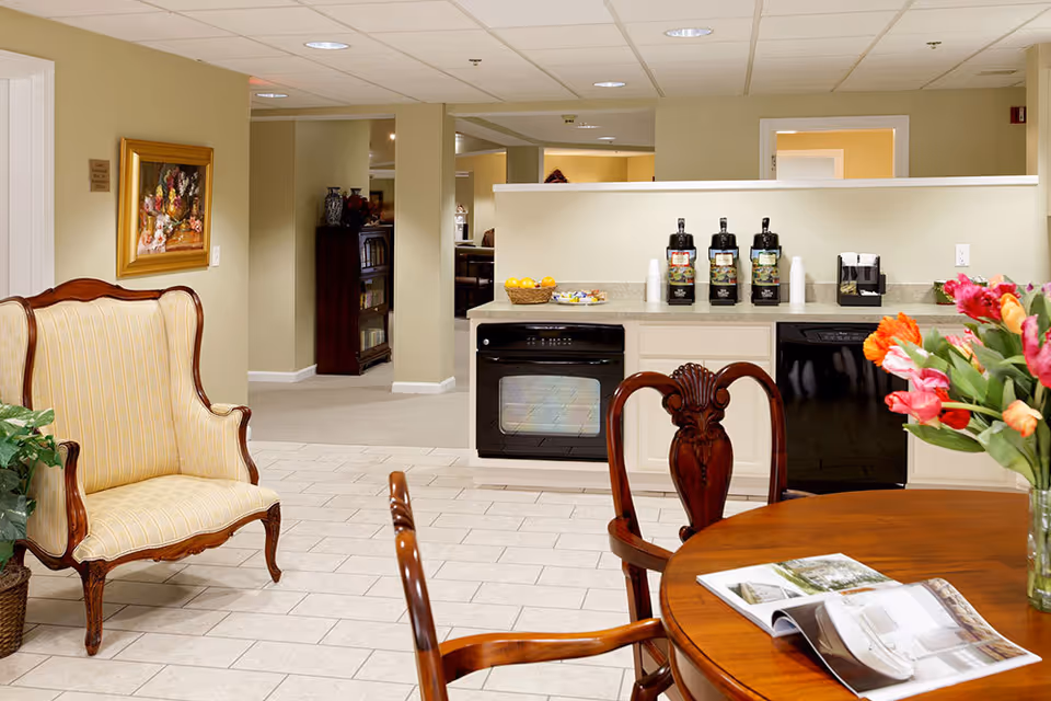 Bright communal dining area with a wooden table and chairs, an upholstered armchair, and a kitchenette coffee station in the background.