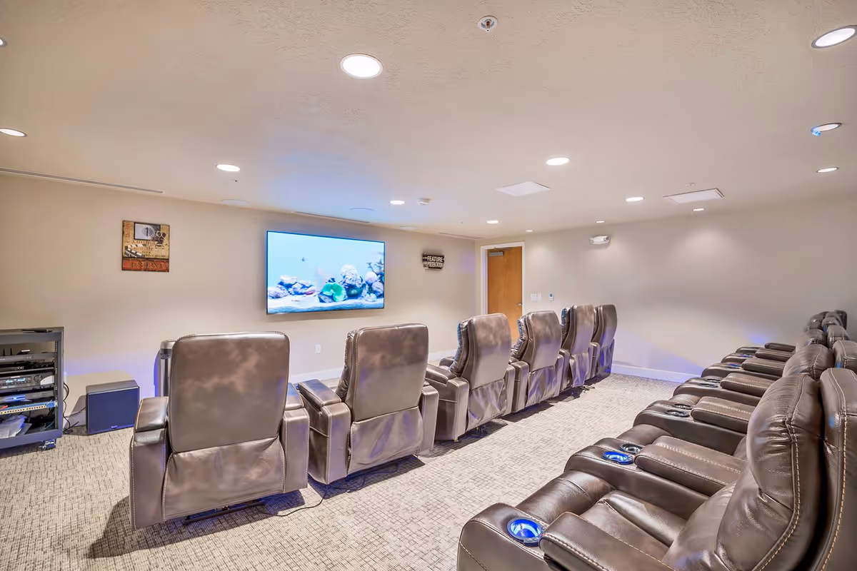 A cozy media room with two rows of brown leather recliner chairs facing a large flat-screen TV mounted on a beige wall. The room has recessed ceiling lights and carpeted flooring, with a door and some wall decorations visible in the background.