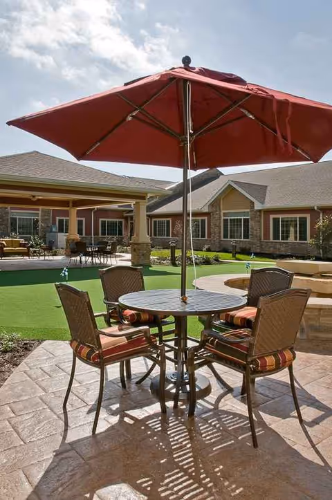 Outdoor patio at a senior living facility with round table, four chairs, and a red umbrella.