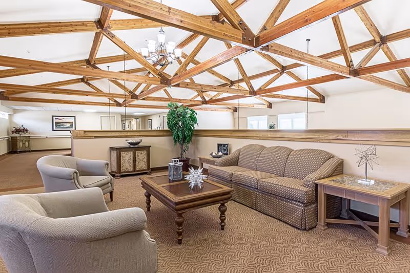 A bright and spacious living room area with exposed wooden ceiling beams, a patterned brown sofa, two beige armchairs, a wooden coffee table with decorative items, a side table, and a potted plant. The room has beige walls and carpeted flooring.