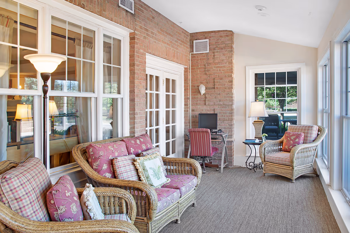 A bright and cozy sunroom with wicker furniture including a loveseat and two armchairs with floral and plaid cushions. There is a floor lamp, a small side table with a lamp and books, and a small desk with a computer and a red striped chair. The room has large windows letting in natural light and a brick accent wall with white trim around the windows and doors.