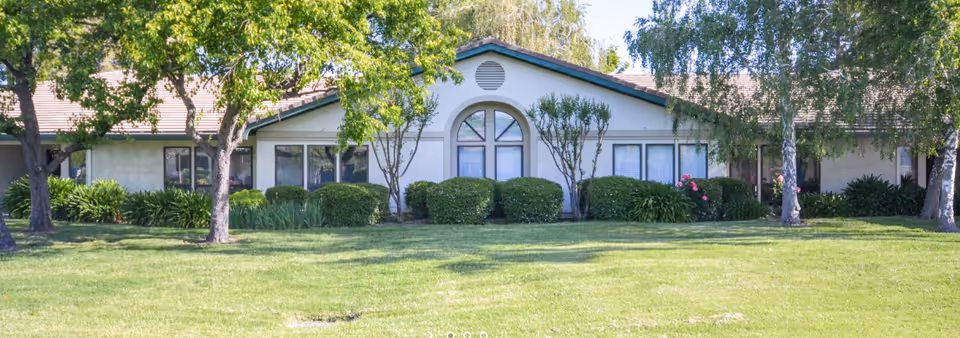 Single-story building front with an arched central window, shrubs and trees on a well-kept lawn.