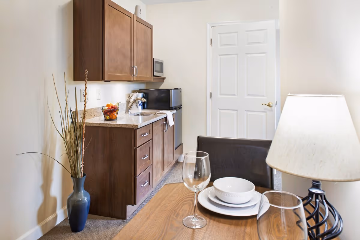 A small kitchenette area with wooden cabinets, a countertop with a sink, a microwave, and a mini refrigerator. A dining table is set with plates, bowls, and wine glasses, next to a lamp with a white shade. A blue vase with tall dried plants is placed on the floor beside the kitchenette. A white door is visible in the background.