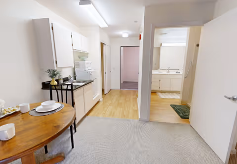 Small studio interior showing a kitchenette with sink and microwave, a round dining table, and an open doorway to a bathroom/vanity area.