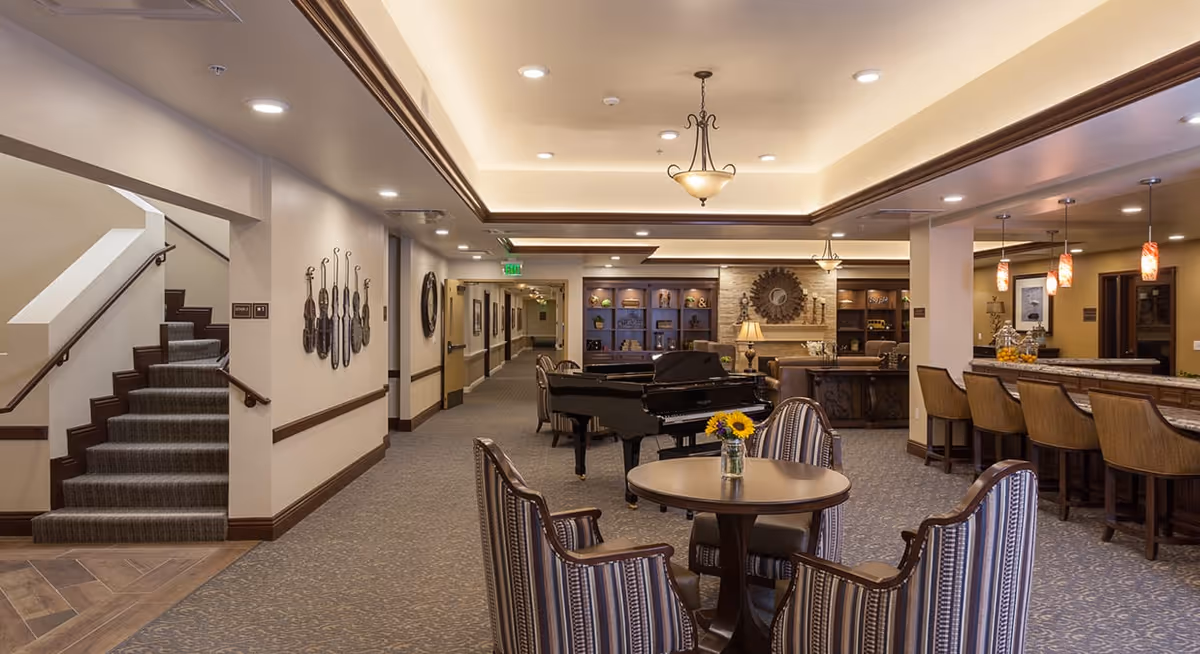 Interior view of a senior living facility common area featuring a round table with four striped upholstered chairs, a grand piano, a bar counter with high chairs, decorative wall art, and warm lighting fixtures. A carpeted staircase is visible on the left side, and the space has a cozy, inviting atmosphere.
