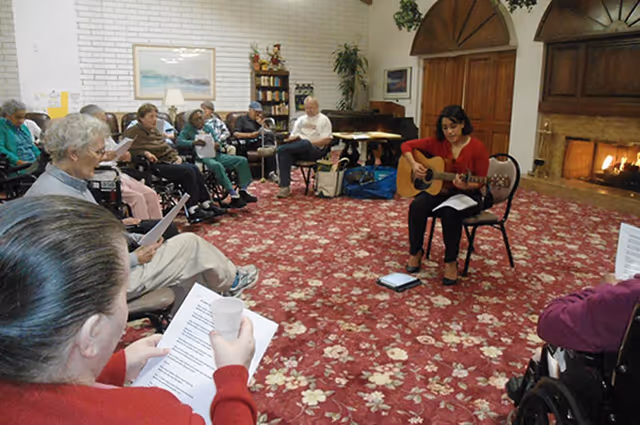 A woman playing guitar leads a sing-along for elderly residents seated in a carpeted common room with a fireplace.