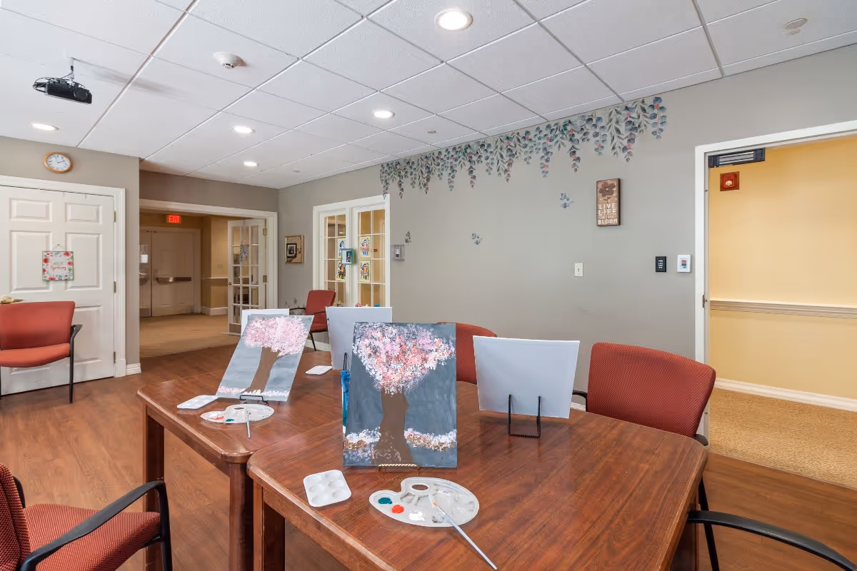 A senior living activity room with tables and chairs set up for painting, featuring canvases of cherry blossom trees.