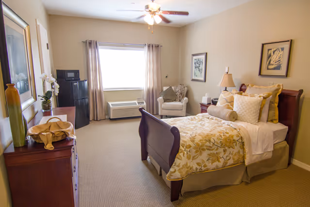 A cozy bedroom with a wooden bed frame, floral bedding, multiple pillows, a nightstand with a lamp, an armchair near the window with curtains, a dresser with decorative items, and a small refrigerator in the corner.