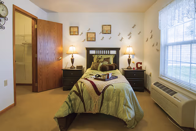 A neatly made single bed with green and brown bedding is centered in a cozy bedroom. Two dark wooden nightstands with matching lamps flank the bed. The walls are decorated with framed art and metal dragonfly sculptures. A window with blinds is on the right side, and an open wooden door on the left reveals a closet with wire shelving.