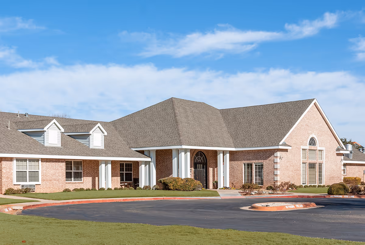 Exterior view of Cimarron Place facility showing a large brick building with a gray shingled roof, multiple windows, and a covered entrance with white columns. The building is surrounded by a well-maintained lawn and a paved driveway with a fire lane marked in red.