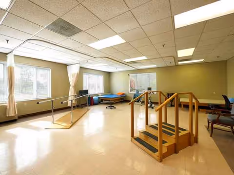 Well-lit therapy room with parallel bars, wooden stairs, and exercise equipment in a rehab facility.