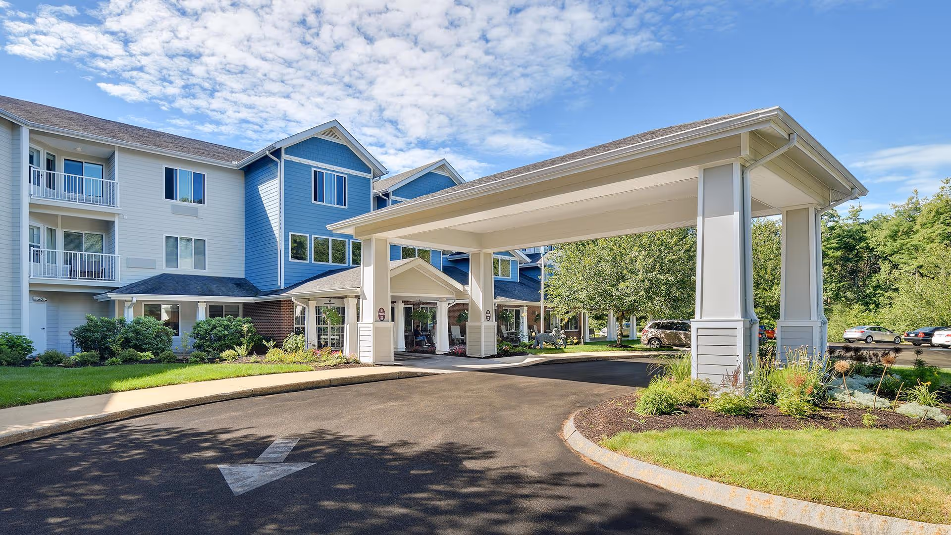 Exterior view of Holiday Maple Suites, a multi-story senior living facility with a covered entrance driveway, landscaped greenery, and parked cars under a partly cloudy sky.