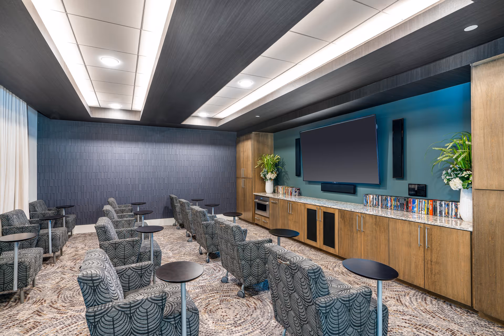 A small theater-style lounge with rows of patterned armchairs and side tables facing a wall-mounted TV above wood cabinetry.