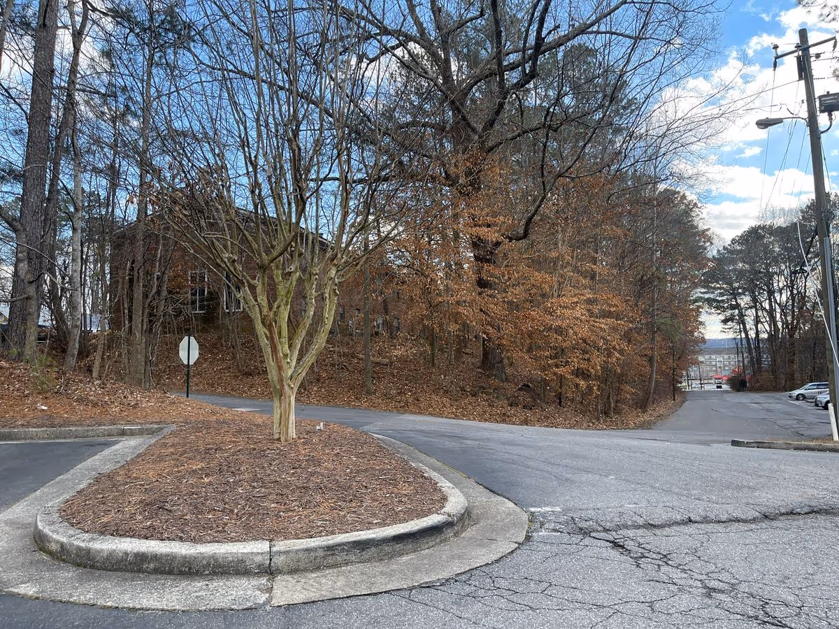 Parking lot entrance with a small tree island, a wooded slope and a partially visible brick building under a partly cloudy sky.