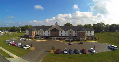Wide exterior view of a large, two-story assisted living facility building named Windsor Estates Assisted Living, surrounded by green lawns and a parking lot with several cars parked. The sky is partly cloudy with blue patches visible.