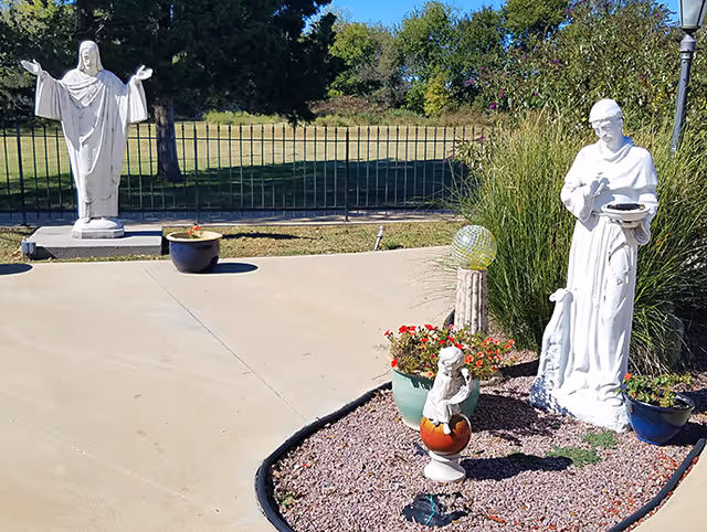 Outdoor paved courtyard with multiple white religious statues, potted flowers, gravel bed and a grassy area beyond a metal fence.