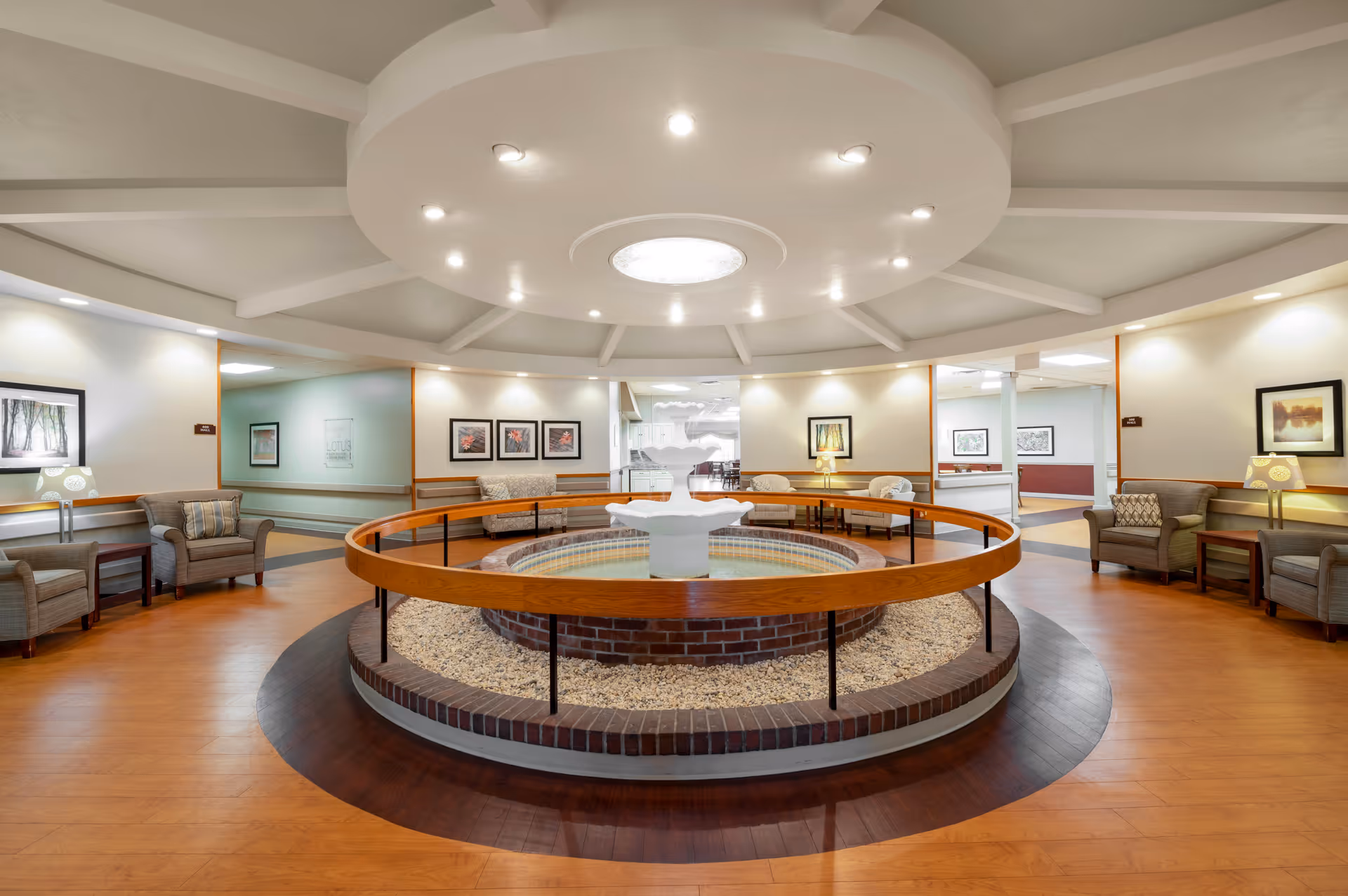 A spacious, well-lit common area with a circular water fountain in the center surrounded by a wooden railing. The room features comfortable armchairs arranged along the walls, framed artwork, and soft lighting from ceiling fixtures and table lamps. The ceiling has a decorative circular design with recessed lights.