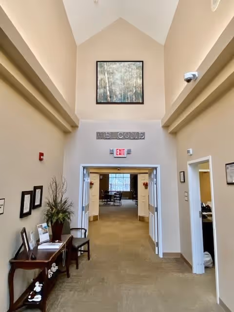 Interior hallway of a senior living facility with beige walls and carpeted floor. A wooden table with chairs and decorative items is on the left side. Above the double doors is a sign that says 'WELCOME' and an exit sign. A framed painting hangs high on the wall above the doorway. The hallway leads to a room with chairs and a window visible in the background.