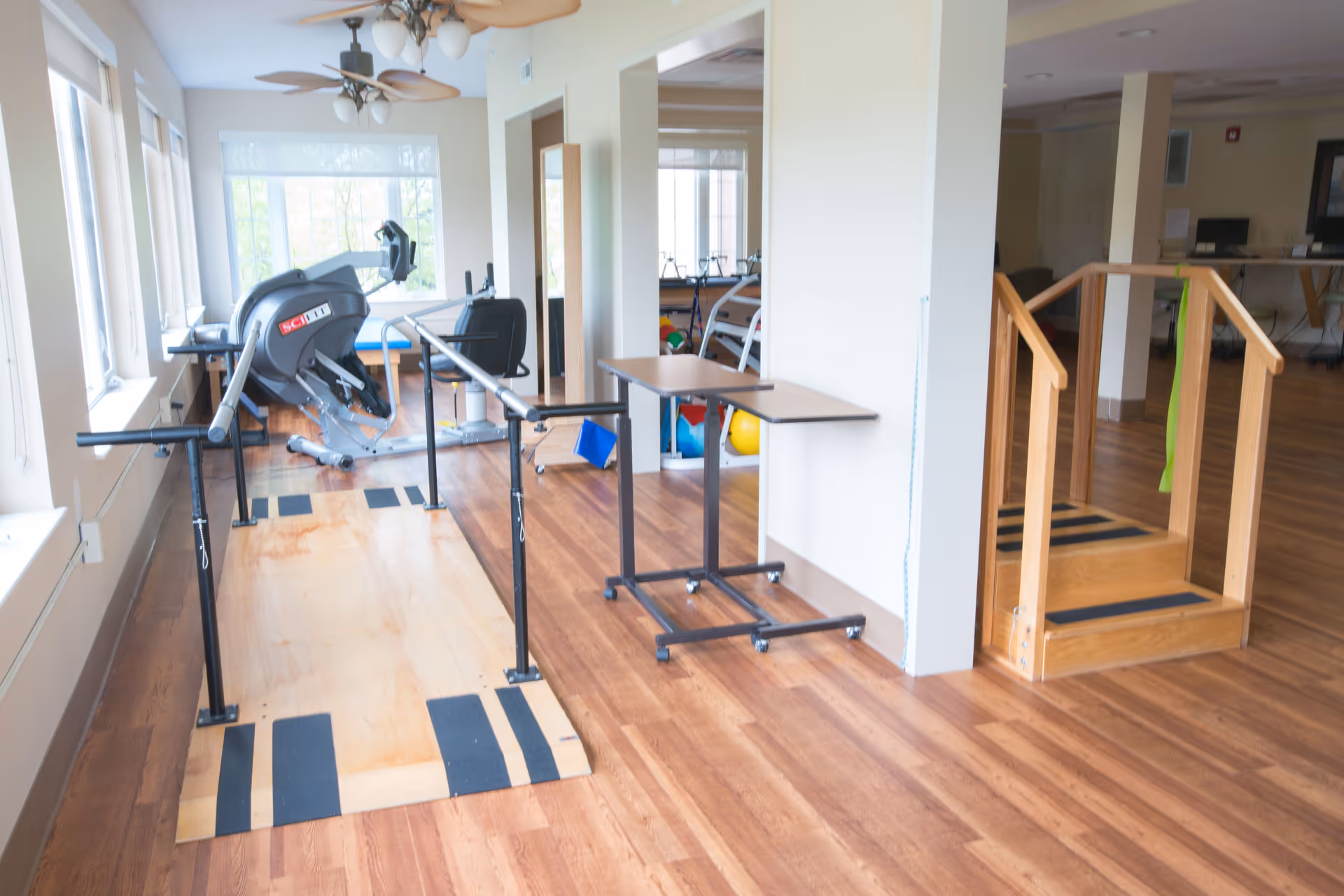 A rehabilitation or physical therapy room with wooden flooring, parallel bars for walking exercises, a small wooden staircase with handrails, exercise equipment including a stationary bike, and a table on wheels. The room has large windows letting in natural light.